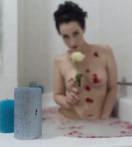 Lydia kniet in der Badewanne in milchigem Wasser, in welchem rote Rosenblütenblätter schwimmen. Sie ist nur verschwommen zu erkennen, blaue Kerzen im Vordergrund sind scharf. Die Blütenblätter haften auch an ihrem nackten Körper. In der rechten Hand hält sie eine weiße Rose und schaut darüber hinweg in die Kamera.