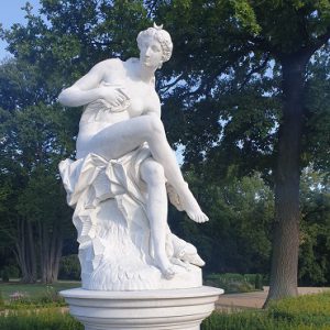Zu sehen ist eine Statue der Luna aus der griechischen Mythologie, die im Park Sanssouci in Potsdam steht. Sie ist aus weißem Stein, sitzt, ein Bein über das andere geschlagen. Der Blick geht zur Seite. Ihren Kopf schmückt ein liegender Halbmond.