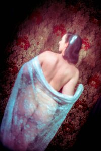 Lydia leicht verschwommen mit dem Rücken zur Kamera stehend vor einem rot-gold gemusterten Vorhang. Sie ist nackt, unterhalb ihrer Schultern hängt ein weiß-transparenter Spitzenstoff, den sie mit beiden Händen festhält.