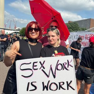 Man sieht zwei Teilnehmer*innen der Tanz-Demo des Berufsverbandes erotische und sexuelle Dienstleistungen in Berlin. Hinter ihnen ist ein aufgespannter, roter Regenschirm zu sehen. Beide halten gemeinsam ein Plakat hoch auf dem zu lesen ist: "Sex work is work".